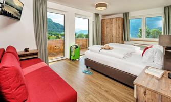A modern hotel room with a large bed and a red sofa. The windows offer a view of the mountains and nature.