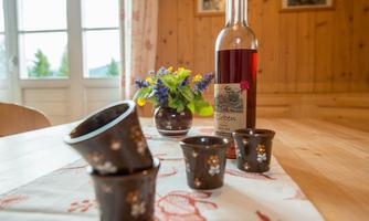 A wooden table with four small cups and a bottle of syrup in a rustic setting. A vase with colorful flowers is also on the table.