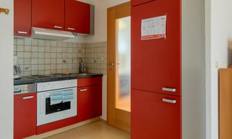 A modern kitchen with red cabinets and a stove. The walls are tiled and there is a door that leads to another room.