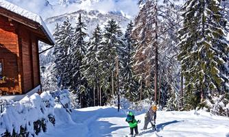 Two children are playing in the snow in front of a wooden house. In the background, there are snow-covered mountains and tall, green trees.