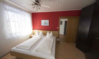 A cozy bedroom with a double bed and red walls. The room features large windows and a ceiling fan.