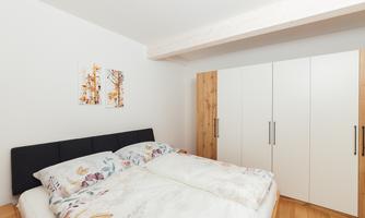 A cozy bedroom with a double bed and colorful bedspreads. In the background, there is a light wardrobe and artworks on the wall.