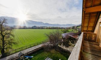 A beautiful view from a balcony over a wide, green meadow and the mountains in the background. The sun shines through the clouds, creating a peaceful atmosphere.