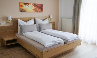 A bright bedroom with a wooden double bed and light bed covers. A colorful picture hangs on the wall, and a large window provides plenty of daylight.