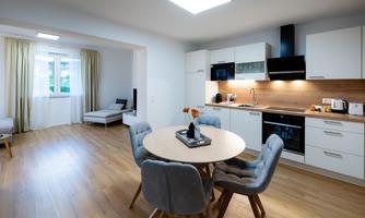 A modern, bright kitchen with a round dining table and comfortable chairs. The room has large windows that let in plenty of light.