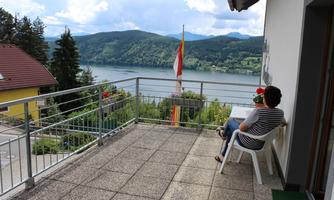 A beautiful balcony with a view of a lake and the surrounding mountains. A person sits relaxed in a chair and enjoys the view.