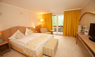 A bright hotel room with a double bed, a cozy armchair, and a balcony. The walls are painted in warm colors and large windows let in plenty of daylight.