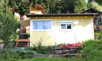A small yellow holiday home surrounded by green meadows and trees. In the foreground, some plants and a wooden walkway are visible.