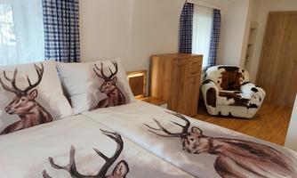 A cozy bedroom with a large bed and bedding featuring a deer motif. Next to the bed is a wooden piece of furniture and an armchair made of cowhide.