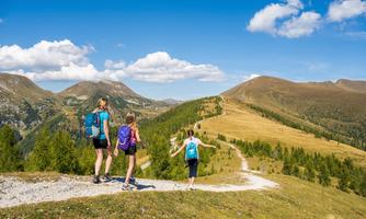 Three hikers enjoy the view of the mountains. The path winds through a green hilly landscape under a clear blue sky.