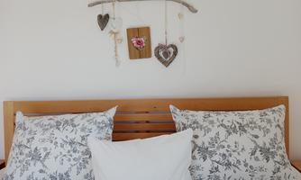 A cozy bed with floral pillows and a white blanket. Above the headboard hang decorative elements made of wood and hearts.
