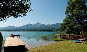 A tranquil lake with a wooden pier and mountains in the background. The surroundings are green and sunny, ideal for relaxing.