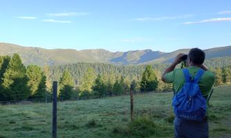 A man with a blue backpack is photographing a beautiful landscape. In the background, green mountains and trees can be seen.