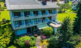 A picturesque hotel building with several floors and a beautiful garden. The green surroundings and sunlight give the scene an inviting atmosphere.