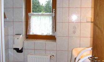 A bright bathroom with white tiles and a window. It includes a toilet, a towel rack, and a shower.