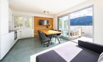 A modern living room with a large dining table and comfortable chairs. Through the large windows, one can see a beautiful view of the lake and the mountains.