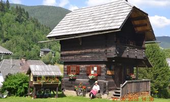 A traditional wooden house in a picturesque landscape, surrounded by trees and green meadows. In front of the house, a person sits in a chair.