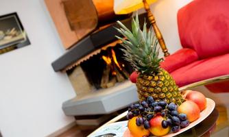 A fresh pineapple and a selection of fruits like grapes and apples on a table. In the background, a cozy fireplace and a red sofa can be seen.