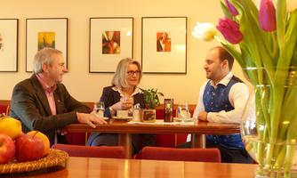 Three people are sitting in a cozy room and having a conversation. On the table are drinks and a fruit bowl.
