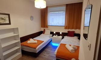 A cozy room with two single beds, light wooden floors, and orange accents. There is a shelf and a window with curtains that let in plenty of light.