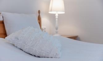 A cozy bedroom with a white double bed and a decorative pillow. A lamp provides soft light.