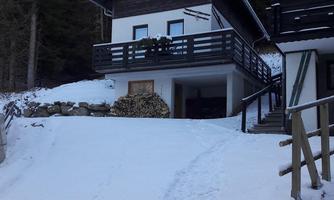 A cozy house in the snow, surrounded by tall fir trees. The balcony is adorned with green decoration.