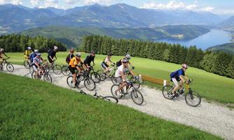 A group of mountain bikers rides along a path in a picturesque landscape. In the background, mountains and a lake can be seen, surrounded by lush greenery.