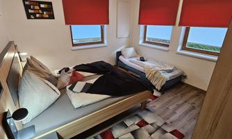 A room with two beds, a large window, and red blinds. The floor is decorated with a modern carpet.