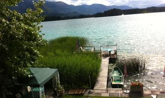 A picturesque lake with green shore plants and a wooden walkway. In the background, gentle mountains and a cloudy sky can be seen.