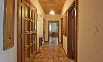 A bright hallway with tiled floors and wooden doors. At the end of the hallway, a table and chairs are visible.