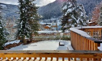 A picturesque winter landscape with snow-covered trees and mountains in the background. In the foreground, a wooden railing can be seen.
