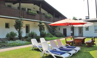 An inviting outdoor area with loungers and a sun umbrella. In the background, there is a building with a garden and lush greenery.