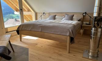 A modern bedroom with a large wooden bed and cozy bedding. Large windows offer a view of the landscape and let in plenty of light.