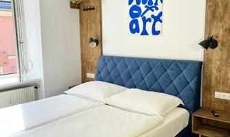 A modern bedroom with a blue bed and a motivational mural. Plants and wooden shelves create a cozy atmosphere.