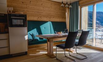 A modern kitchen with wooden paneling and a large table in the center. There are chairs and large windows that provide plenty of light and a view of nature.