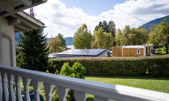 A view from a balcony over a green meadow with a solar panel and some wooden accommodations. In the background, trees and hills are visible.
