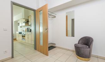 A modern hallway with an open-plan kitchen in the background. On the right, there is an elegant chair and a mirror on the wall.