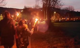 Eine Gruppe von Menschen geht mit Fackeln entlang einem Weg. Der Himmel wird von der Dämmerung erhellt, während im Hintergrund weitere Lichter zu sehen sind.