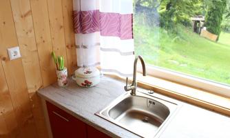 A modern kitchen with a stainless steel sink and a window that looks out onto a green meadow. The walls are wooden-paneled and combined with a colorful curtain.