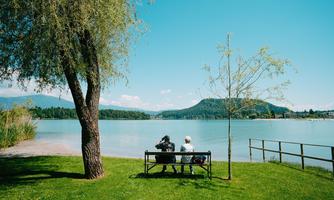 A calm lake with green grass and gentle hills in the background. Two people are sitting on a bench enjoying the view.