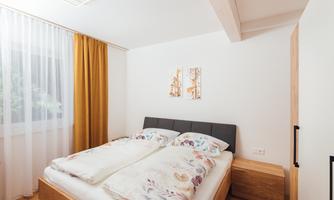 A bright bedroom with a large double bed and colorful bedspreads. The windows have light curtains and there are wooden shelves and pictures on the wall.