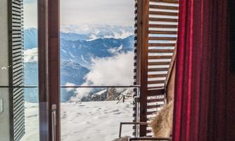 A modern apartment with a large glass front and a view of snow-covered mountains. On the balcony, there is a chair and a fur that invites you to relax.