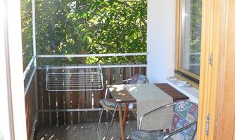 A small balcony with a table and chairs. In the background, there is a lot of greenery from trees visible.