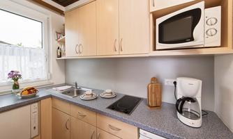A modern kitchen with light wooden cabinets and gray countertops. It is well equipped with a microwave, kettle, and dishes.