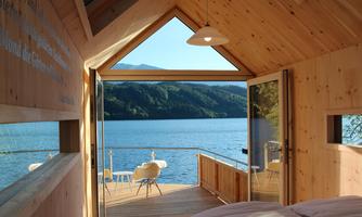 A cozy wooden house with a view of a clear lake and green mountains. Large windows let in plenty of daylight.