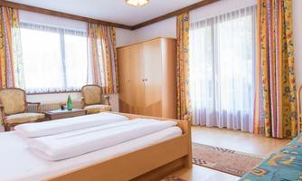 A bright hotel room with a double bed, a TV, and comfortable seating. The large windows provide plenty of daylight and a view outside.