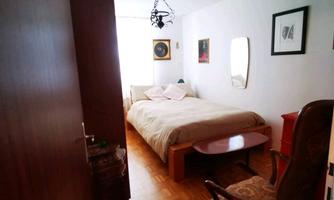 A simple bedroom with a bed, a chair, and subtle wall pictures. The room has wooden flooring and plenty of daylight.
