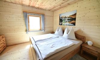 A bright bedroom with wooden paneling and a large window. The bed is cozy, decorated with two pillow-like cushions.