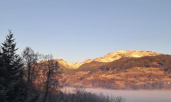 A picturesque mountain landscape under a clear sky. In the foreground, trees can be seen, and a gentle mist lies on the slope.