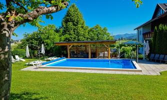 A beautiful garden with a clear pool and sunny loungers. In the background, you can see trees and a roof over the seating area.